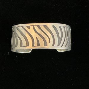 May include: A wide silver cuff bracelet with a repeating wave pattern. The design features alternating polished and etched lines, creating a textured effect. The bracelet has a simple, open design.