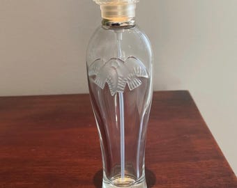 May include: A clear glass perfume bottle with a tall, slender body and a frosted glass stopper featuring an intricate swirl pattern. The front of the bottle displays a frosted glass relief of three birds in flight, with a silver collar and white tube.