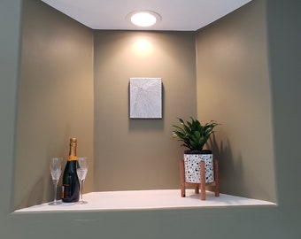 Könnte beinhalten: Ein weißes, strukturiertes Leinwandbild hängt an einer Wand in einer Nische mit einer Topfpflanze und einer Flasche Champagner mit zwei Champagnergläsern auf einer weißen Oberfläche.