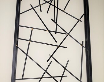 Könnte beinhalten: Eine schwarze Wandskulptur aus Metall mit einem rechteckigen Rahmen und einem geometrischen Muster aus sich kreuzenden Linien. Die Linien bestehen aus dünnen schwarzen Stäben und sind mit goldfarbenen Befestigungselementen am Rahmen befestigt.