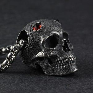 May include: A silver skull pendant with a red gemstone set in the forehead. The skull is detailed with a textured surface and a chain is attached to the back.