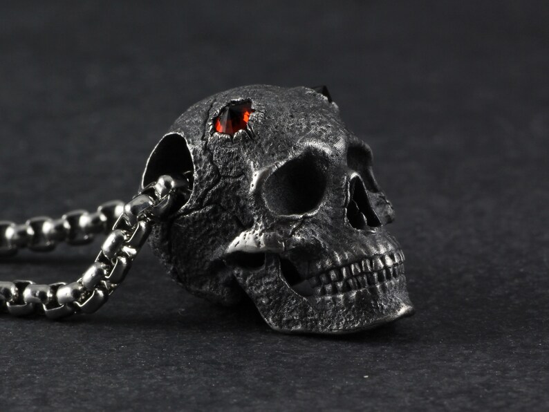 May include: A silver skull pendant with a red gemstone set in the forehead. The skull is detailed with a textured surface and a chain is attached to the back.