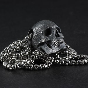 May include: A silver skull pendant necklace with a detailed, textured skull design. The necklace features a silver chain.