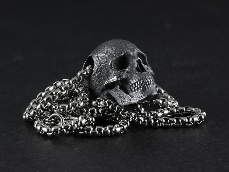 May include: A silver skull pendant necklace with a detailed, textured skull design. The necklace features a silver chain.