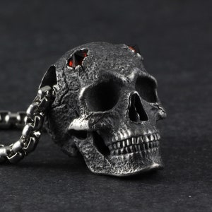 May include: A silver skull pendant with red gemstone eyes, attached to a silver chain. The skull has a textured surface and detailed features.