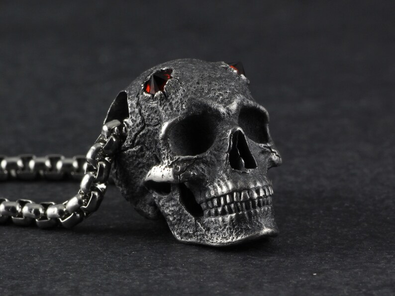 May include: A silver skull pendant with red gemstone eyes, attached to a silver chain. The skull has a textured surface and detailed features.