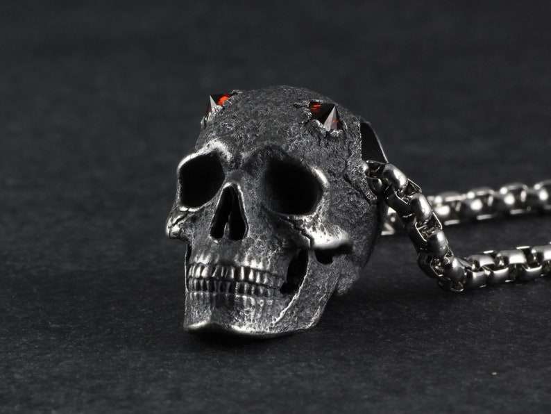 May include: A silver skull pendant with red gemstone eyes and horns, attached to a silver chain.