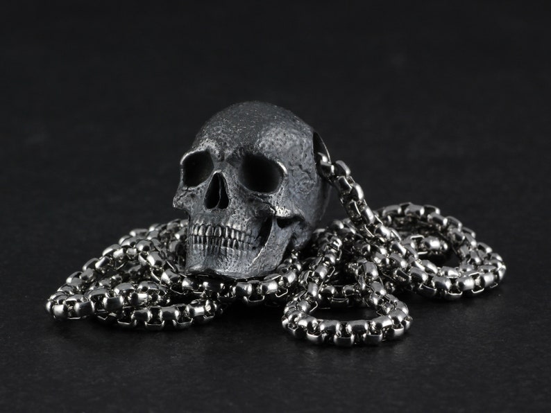 May include: A silver skull pendant on a silver chain necklace. The skull is detailed with a textured surface and has a realistic appearance.