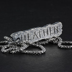 May include: A silver necklace with a rectangular pendant that says "HEATHEN" in a rustic font.