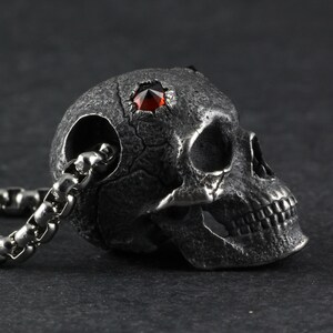 May include: A silver skull pendant with a red gemstone set in the forehead. The skull is detailed with a textured surface and a chain is attached to the back.