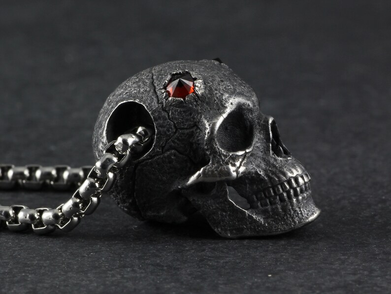 May include: A silver skull pendant with a red gemstone set in the forehead. The skull is detailed with a textured surface and a chain is attached to the back.
