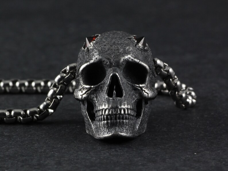 May include: A silver skull pendant with red gemstone eyes and horns, attached to a silver chain.