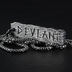 May include: A silver necklace with a pendant that says "DEVILANT" in a stylised font. The pendant is made to look like a piece of wood with a rough texture.