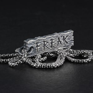 May include: A silver necklace with a rectangular pendant that says "FREAK" in a textured, wood-like style.