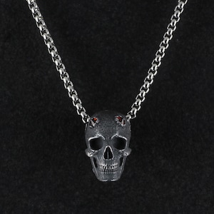 May include: A silver skull pendant necklace with red gemstone eyes. The pendant has a detailed design and is attached to a silver chain.