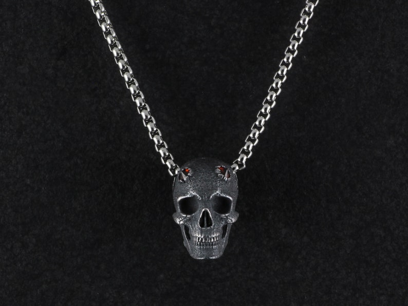 May include: A silver skull pendant necklace with red gemstone eyes. The pendant has a detailed design and is attached to a silver chain.