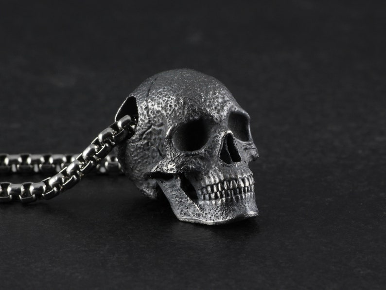 May include: A silver skull pendant necklace with a detailed, textured design. The skull has a chain link necklace.