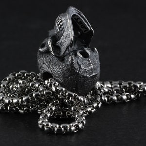 May include: A silver skull pendant with a detailed design, hanging on a silver chain. The skull is open at the jaw, revealing the inside.