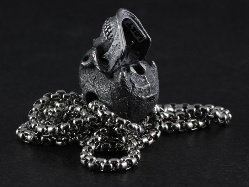 May include: A silver skull pendant with a detailed design, hanging on a silver chain. The skull is open at the jaw, revealing the inside.