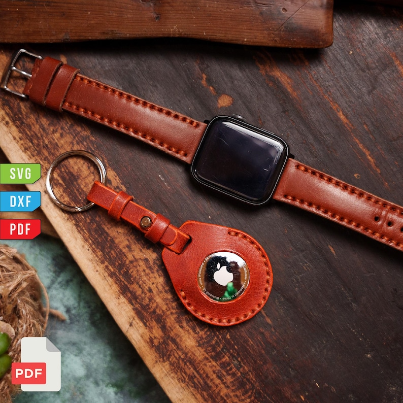 Pu&ograve; includere: Un cinturino in pelle marrone e uno smartwatch nero sono esposti accanto a un portachiavi in pelle marrone abbinato con un anello argentato. Il portachiavi contiene un Apple AirTag argentato. Gli oggetti sono su una superficie di legno.