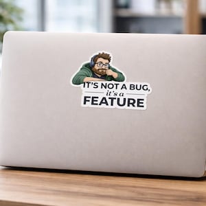 以下が含まれることがあります： ラップトップに貼られたステッカーには、ヘッドホンとメガネをかけた人物の漫画の画像が描かれています。ステッカーには「IT'S NOT A BUG, it's a FEATURE」というテキストが白黒で表示されています。