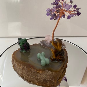 May include: A crystal tree with purple amethyst stones on a brown agate slice. A green jade cat figurine and a brown tiger eye dog figurine are on the slice. A green jade bone is in front of the dog.
