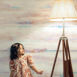 May include: A young girl in a floral print dress stands in front of a wall with a watercolor-like design. She is looking up at a tripod floor lamp with a white lampshade.