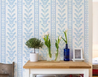 May include: A room with light blue and white floral wallpaper. A white table with a wooden top holds a potted plant, vases with flowers, and a blue bottle. A wooden chair is to the left.