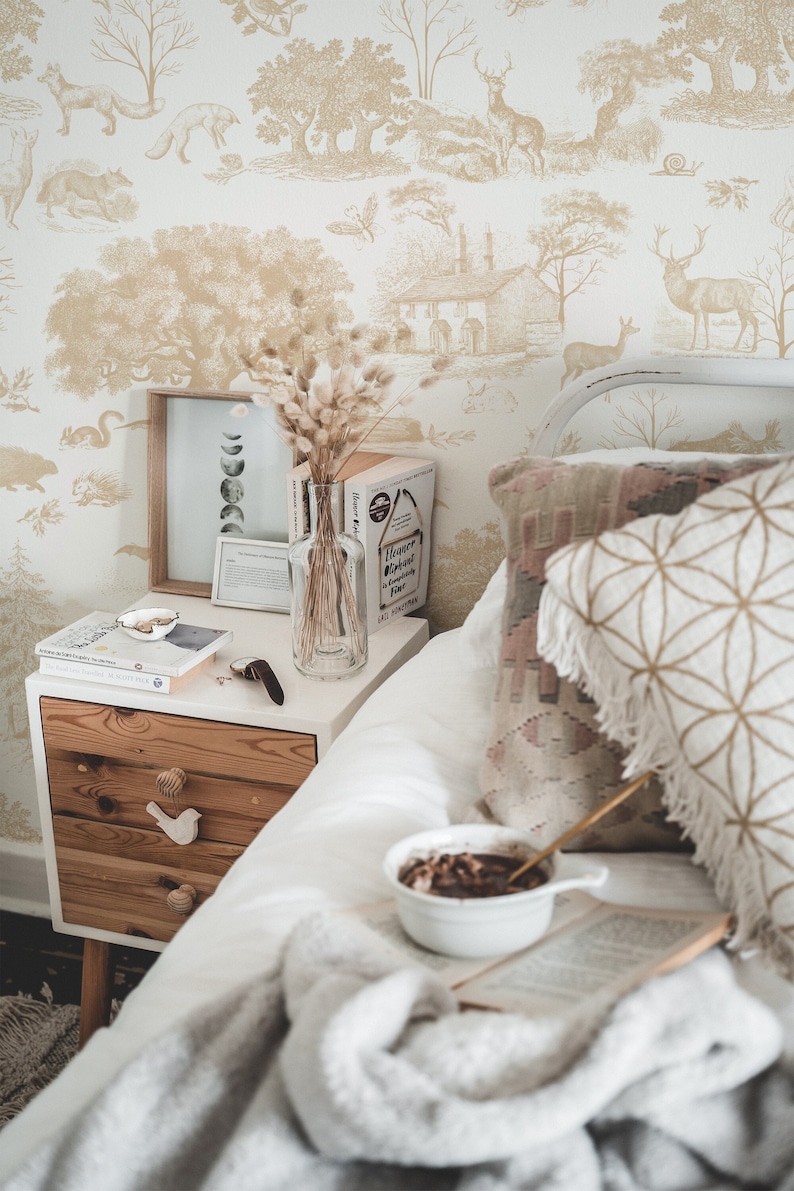 May include: A cozy bedroom scene featuring a white nightstand with wooden drawers, adorned with a vase of dried flowers, books, and a framed print. A bowl of cereal and a book rest on a soft blanket on the bed. The wallpaper has a beige forest scene.