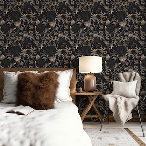 K&ouml;nnte beinhalten: Ein Schlafzimmer-Interieur mit einem wei&szlig;en Bett, einem braunen Kunstfellkissen und einem wei&szlig;en Stuhl mit einer Wolldecke. Die Wand ist mit schwarzer Tapete mit einem floralen Muster in Gold und Grau bedeckt. Eine Lampe mit einem wei&szlig;en Schirm und einem braunen Fu&szlig; steht auf einem Holz-Beistelltisch.