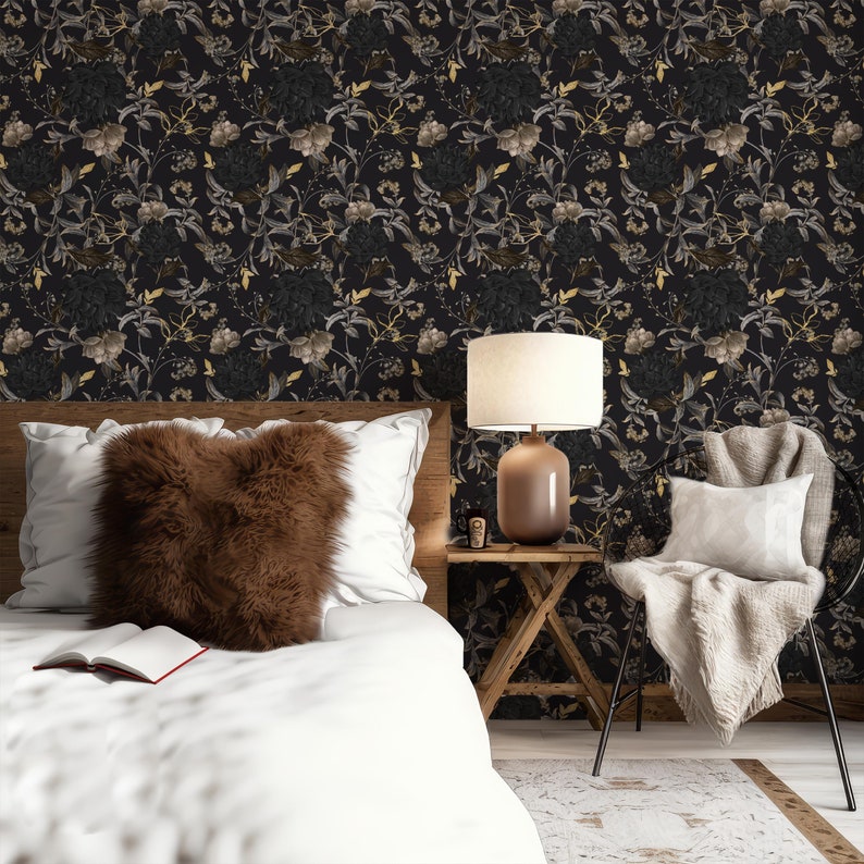 K&ouml;nnte beinhalten: Ein Schlafzimmer-Interieur mit einem wei&szlig;en Bett, einem braunen Kunstfellkissen und einem wei&szlig;en Stuhl mit einer Wolldecke. Die Wand ist mit schwarzer Tapete mit einem floralen Muster in Gold und Grau bedeckt. Eine Lampe mit einem wei&szlig;en Schirm und einem braunen Fu&szlig; steht auf einem Holz-Beistelltisch.