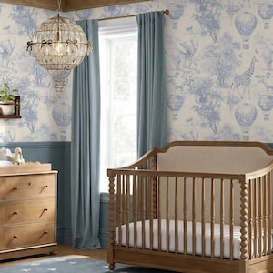 May include: A nursery room featuring a wooden crib with a beige upholstered headboard, a wooden dresser, and a spherical chandelier. The walls are decorated with blue and white toile wallpaper depicting hot air balloons, elephants, and giraffes.