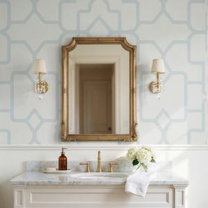 Può includere: Uno specchio con cornice dorata posizionato tra due applique in ottone con paralumi color crema. Sotto, un mobile lavabo in marmo bianco presenta un rubinetto in ottone, un dispenser di sapone marrone, un asciugamano bianco e un vaso di ortensie.