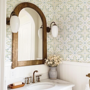 Op de afbeelding: Een badkamer met een houten spiegel, witte wandkandelaars en een marmeren aanrecht met een wastafel en kraan. De muur is versierd met tegels met blauwe en groene bloempatronen. Een vaas met droogbloemen staat op het aanrecht.
