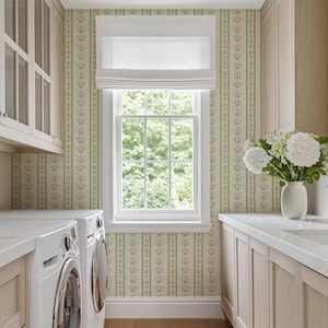 May include: A white washing machine and tumble dryer in a utility room with light wood cabinets and a window with a view of green trees. The walls are covered in a floral wallpaper with green and pink stripes.