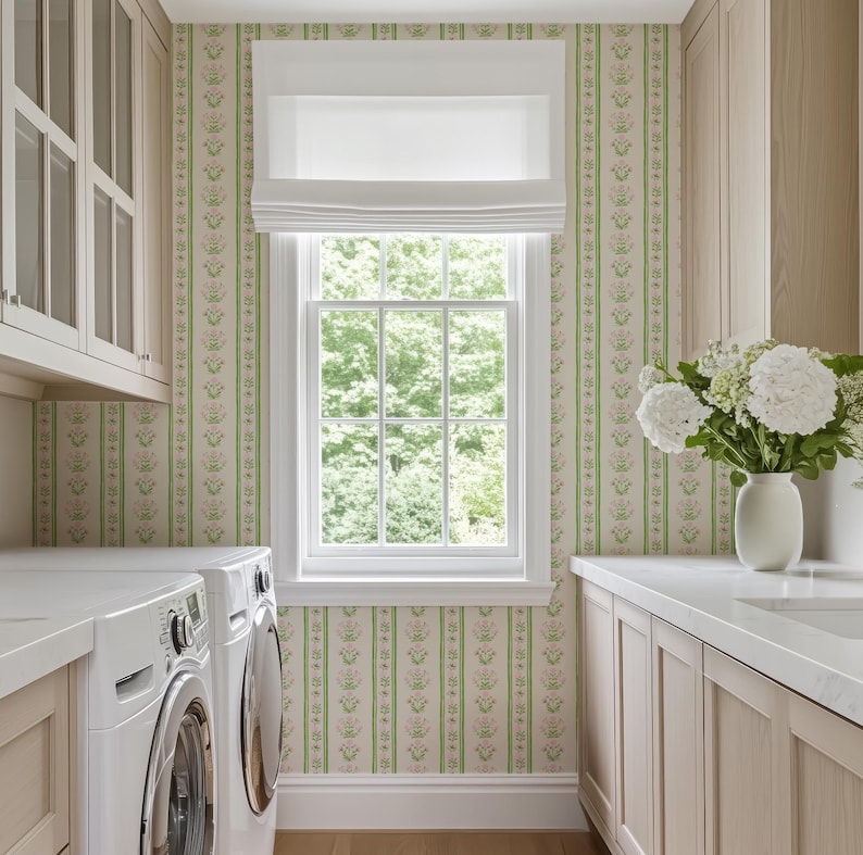 May include: A white washing machine and tumble dryer in a utility room with light wood cabinets and a window with a view of green trees. The walls are covered in a floral wallpaper with green and pink stripes.