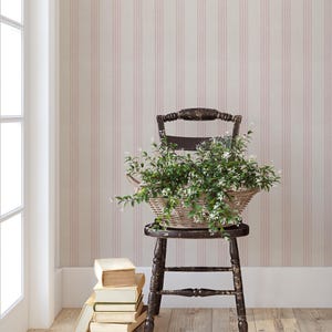 May include: A dark wooden chair supports a wicker basket overflowing with greenery and white flowers. A pile of old books sits on the floor. The backdrop is striped wallpaper in pale pink and white.