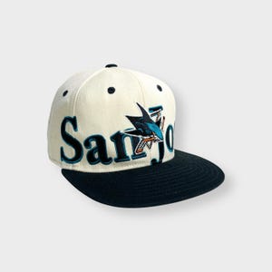 Könnte beinhalten: Eine weiße und schwarze Baseballkappe mit einem blauen und türkisfarbenen Hai-Logo und dem Text "San Jose" auf der Vorderseite.