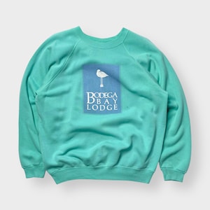 Könnte beinhalten: Türkisfarbenes Sweatshirt mit Rundhalsausschnitt und Raglanärmeln. Ein hellblaues Quadrat auf der Vorderseite zeigt eine weiße Vogelillustration über den Worten "Bodega Bay Lodge" in einer Serifenschrift. Das Sweatshirt ist einfarbig.