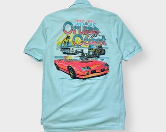 May include: Light blue polo shirt featuring a colorful graphic on the back. The graphic includes illustrations of classic cars and the text "Fourth Annual Santa Cruz Cruise to the Coast". The shirt has short sleeves and a collared neckline.