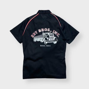 May include: Black short-sleeved work shirt with a white and red design. The back of the shirt features a graphic of a semi-truck with the text "EBY BROS., INC." and "Boise, Idaho". Red and white stripes run down the shoulders.