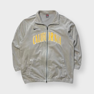 Könnte beinhalten: Hellgraue Trainingsjacke mit Reißverschluss und dem Wort "CALIFORNIA" in gelben Buchstaben auf der Brust. Die Jacke hat ein kleines schwarzes Nike-Logo und schwarze Streifen an den Ärmeln.
