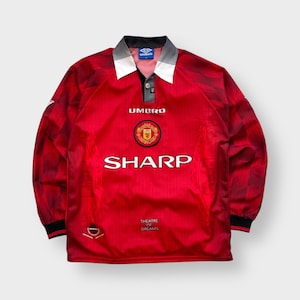 Może przedstawiać: Czerwona koszulka piłkarska z długim rękawem z czarno-biało-szarym kołnierzem. Koszulka zawiera napis "SHARP" w kolorze białym, herb drużyny oraz napis "THEATRE OF DREAMS". Logo Umbro jest widoczne na kołnierzu.