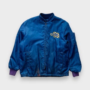 May include: A vintage blue jacket with a zip fastening and a "Monroe" patch on the left side. The jacket has a purple lining and a chequered flag design on the patch.
