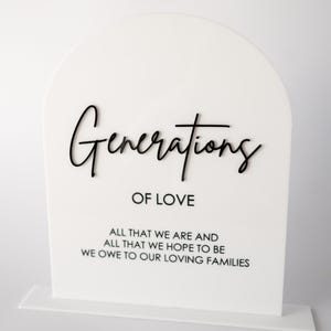 May include: White acrylic sign with a rounded top and a black inscription that reads "Generations of Love" and a quote below that reads "All that we are and all that we hope to be we owe to our loving families."