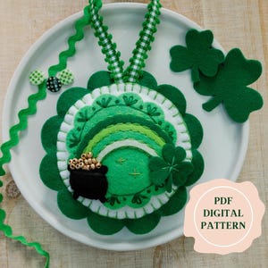 May include: A felt ornament with a green shamrock, a rainbow, and a pot of gold. The ornament is on a white plate with a green and white ribbon. The text "PDF DIGITAL PATTERN" is on a pink circle.