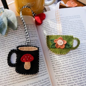May include: Felt bookmarks in the shape of a black coffee mug with a red mushroom and a green teacup with a flower. Both have decorative ribbon ties and are placed on an open book.
