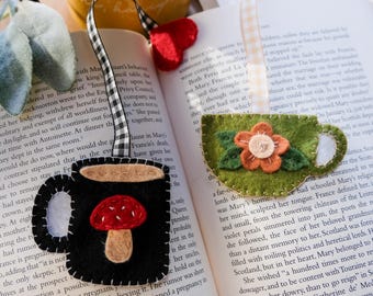 May include: Felt bookmarks in the shape of a black coffee mug with a red mushroom and a green teacup with a flower. Both have decorative ribbon ties and are placed on an open book.