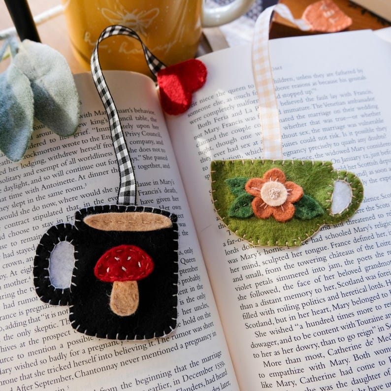 May include: Felt bookmarks in the shape of a black coffee mug with a red mushroom and a green teacup with a flower. Both have decorative ribbon ties and are placed on an open book.