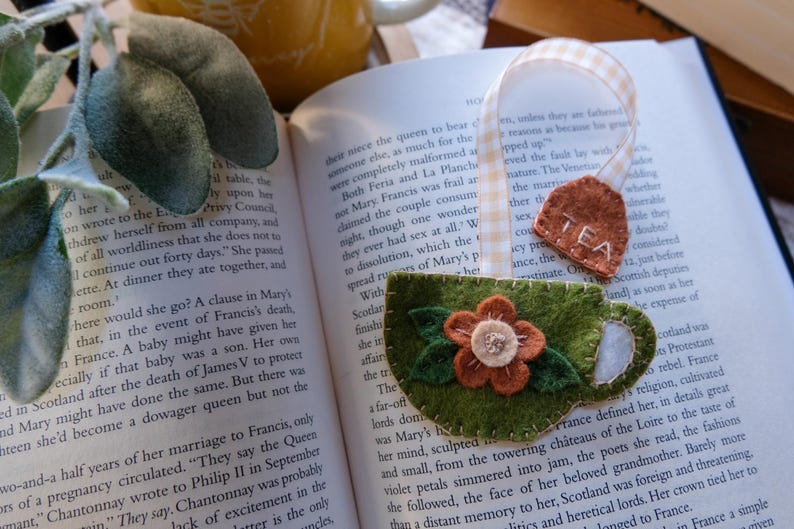 May include: A handmade felt bookmark featuring a green teacup design with an orange and cream flower applique. A small brown tea bag tag with "TEA" stitched on it is attached to a ribbon. The bookmark rests on an open book.